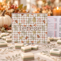 A personalized mahjong set with a clear acrylic frame and a gingham board printed with autumn leaves, pinecones, and the name 'Terese' in brown script. The board is displayed on a patterned tablecloth surrounded by completed mahjong tiles, with a blurred fall-themed background featuring a pumpkin and candles.