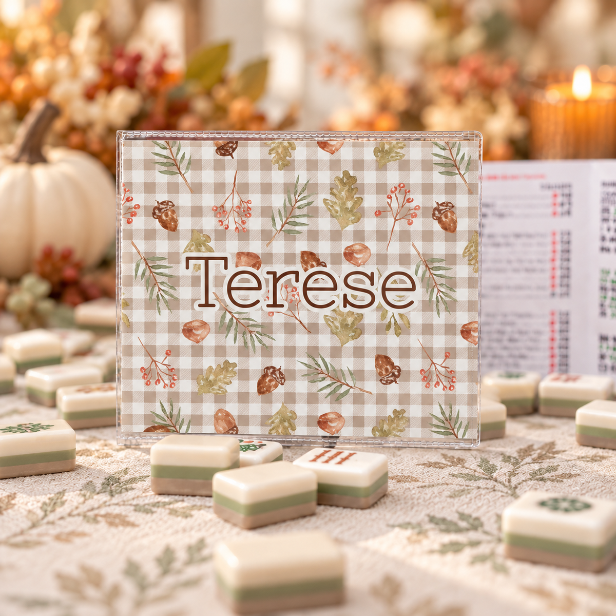 A personalized mahjong set with a clear acrylic frame and a gingham board printed with autumn leaves, pinecones, and the name 'Terese' in brown script. The board is displayed on a patterned tablecloth surrounded by completed mahjong tiles, with a blurred fall-themed background featuring a pumpkin and candles.