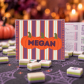 A Halloween-themed game board set with a clear acrylic board featuring orange pumpkins and the name 'MEGAN' on a purple, orange, and cream striped background. The board is surrounded by small, two-toned game pieces on a dark table with a spiderweb print, with a blurred festive background.
