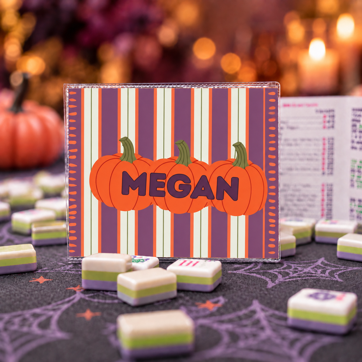 A Halloween-themed game board set with a clear acrylic board featuring orange pumpkins and the name 'MEGAN' on a purple, orange, and cream striped background. The board is surrounded by small, two-toned game pieces on a dark table with a spiderweb print, with a blurred festive background.