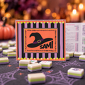 A Halloween-themed card game setup featuring a central card with a witch hat and the name 'SAMI' on an orange background, surrounded by small game pieces on a dark patterned surface with a blurred festive backdrop.