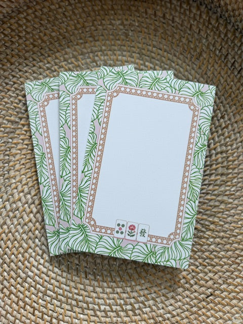 A stack of rectangular notepad sheets with a green tropical leaf border and a tan decorative frame, placed on a woven straw surface.