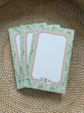 A stack of rectangular notepad sheets with a green tropical leaf border and a tan decorative frame, placed on a woven straw surface.