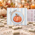 A square decorative card or sign with a watercolor orange pumpkin inside a circular frame on a light blue striped background, with the word 'Savor' in script above it. The sign is styled on a table with small layered tiles and a blurred autumn-themed background.