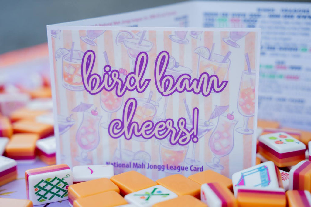 A National Mah Jong League Card reading 'bird bam cheers!' in purple script, surrounded by miniature orange and white Mahjong tiles on a blurred background.